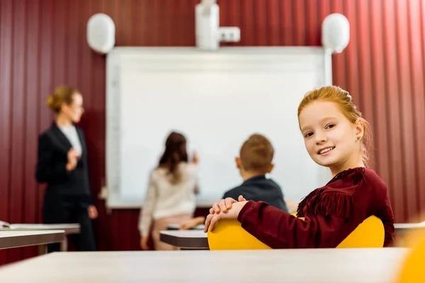 Adorable schoolchild smiling at camera while classmates studying with whiteboard behind — Stock Photo