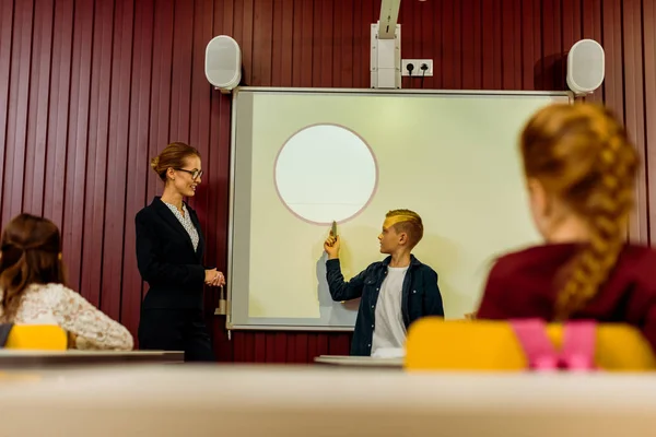 Enseignant et écoliers regardant garçon faisant la présentation au tableau blanc interactif — Photo de stock
