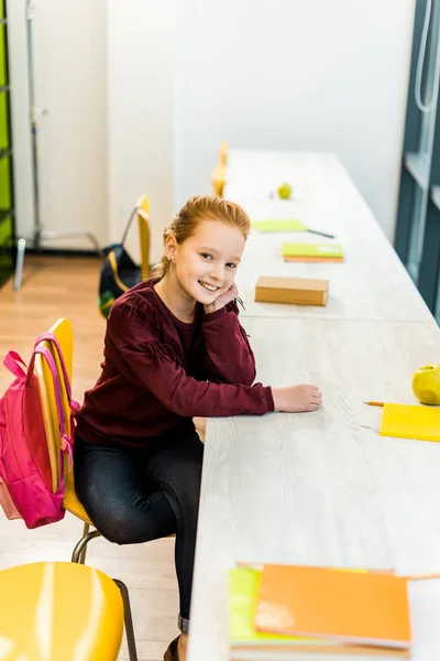 High angle view of beautiful schoolgirl smiling at camera while sitting at desk in library — Stock Photo