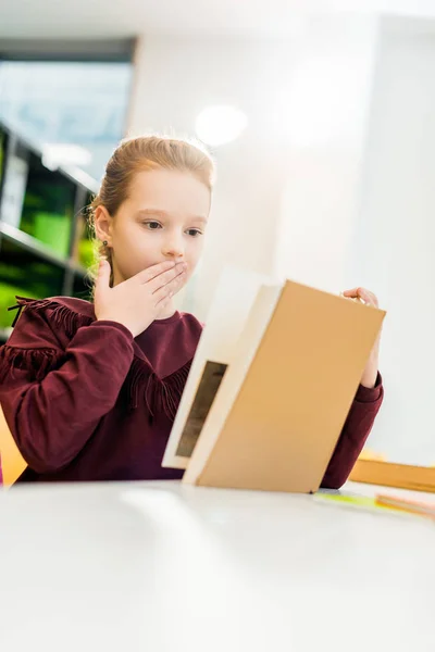 Sorprendido colegiala lectura libro mientras sentado en la biblioteca - foto de stock