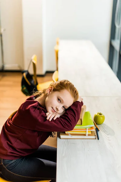 Nettes Schulkind lehnt an Büchern auf dem Schreibtisch und blickt in Bibliothek in die Kamera — Stockfoto