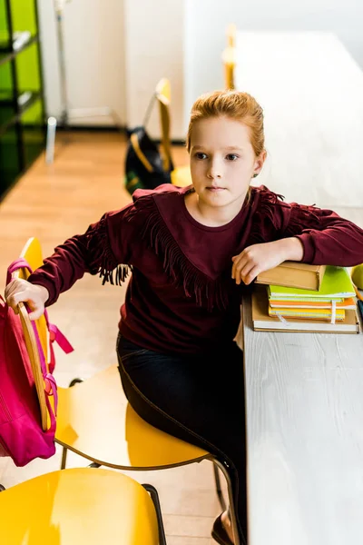 Entzückendes Schulkind, das mit Büchern am Schreibtisch sitzt und in der Bibliothek wegschaut — Stockfoto