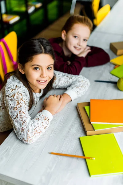 High angle view of adorable schoolgirls leaning at desk with books and smiling at camera — Stock Photo