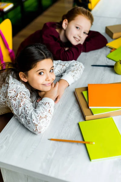 Blick von Schulmädchen, die sich mit Büchern am Schreibtisch lehnen und in die Kamera lächeln — Stockfoto