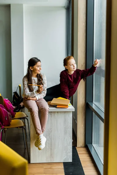 Adorable happy schoolgirls sitting on desk and looking at window — Stock Photo