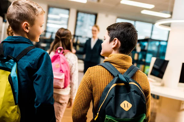 Vista posteriore dei ragazzi con gli zaini che si guardano mentre visitano la biblioteca con i compagni di classe — Foto stock