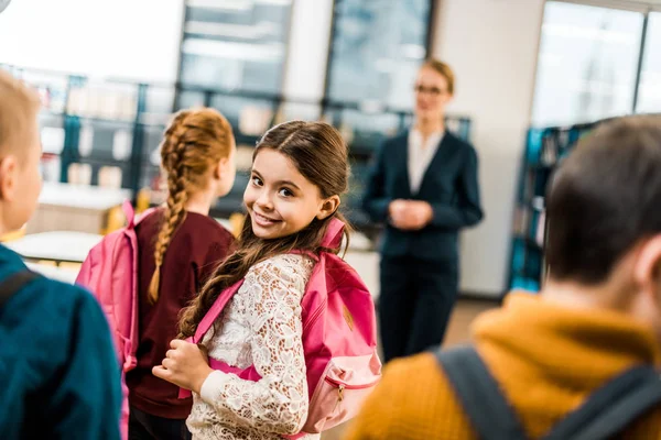 Школьник с рюкзаком, улыбающийся в камеру во время посещения библиотеки с одноклассниками — стоковое фото