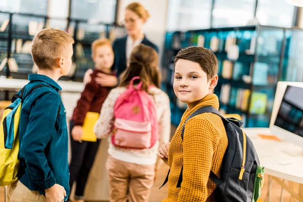 Niño con mochila mirando a la cámara mientras visita la biblioteca con sus compañeros de clase - foto de stock
