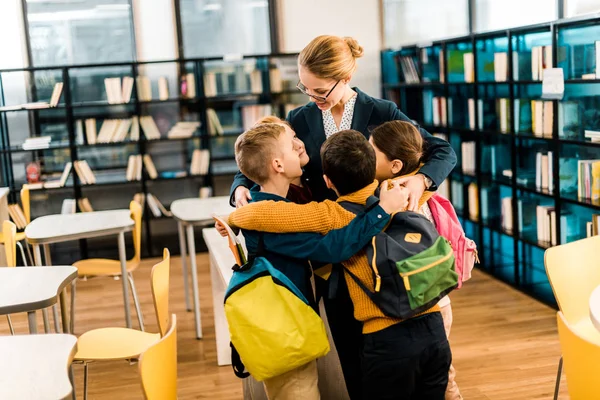 Crianças escolares felizes com mochilas abraçando bibliotecário na biblioteca — Fotografia de Stock