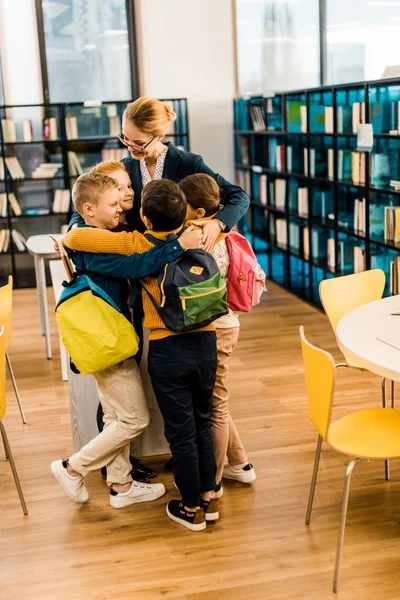 Happy kids with backpacks hugging librarian in library — Stock Photo