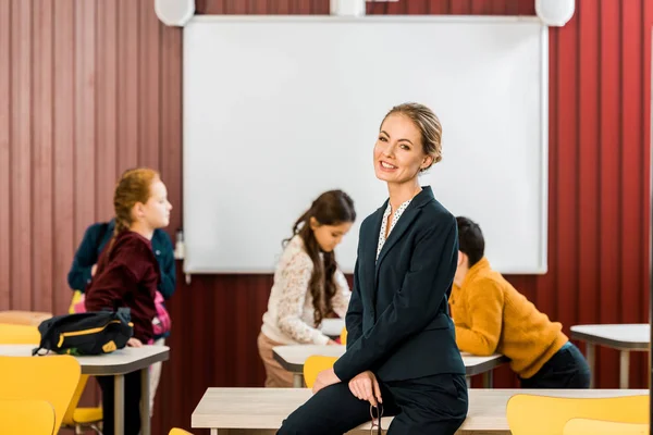 Beautiful young teacher smiling at camera while kids packing backpacks behind — Stock Photo