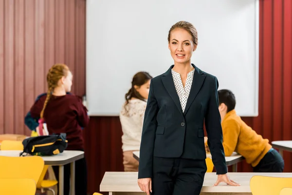 Beautiful young female teacher smiling at camera while schoolkids standing behind near whiteboard — Stock Photo