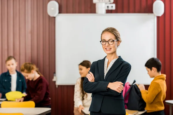 Belle jeune enseignant lunettes debout avec les bras croisés et souriant à la caméra tandis que les enfants emballant des sacs à dos derrière — Photo de stock