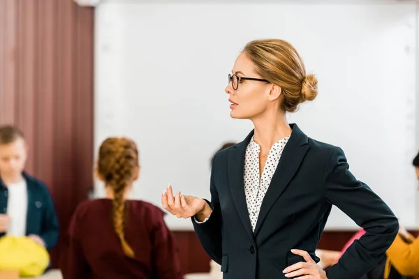 Young female teacher in eyeglasses looking away while schoolkids standing behind — Stock Photo