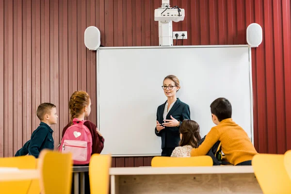 Jeune enseignant debout près du tableau blanc interactif et regardant les écoliers avec des sacs à dos — Photo de stock