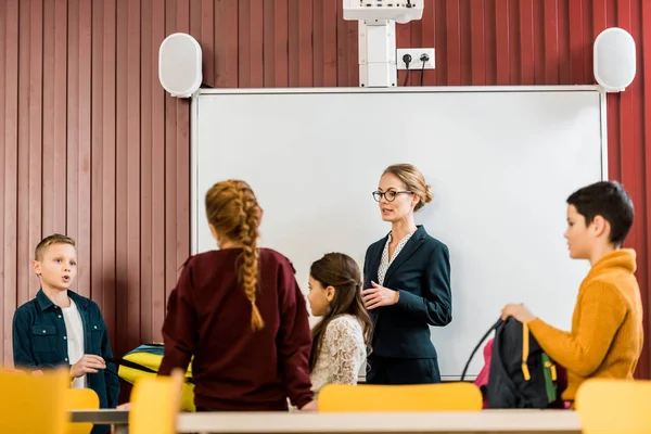 Young teacher standing near interactive whiteboard and looking at schoolchildren with backpacks — Stock Photo