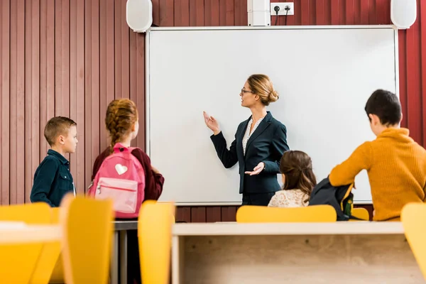 Visão traseira de escolares com mochilas olhando para o professor fazendo apresentação no quadro interativo — Fotografia de Stock