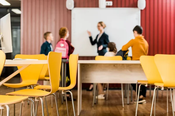Chairs and desks on foreground and kids looking at teacher showing at interactive whiteboard behind — Stock Photo