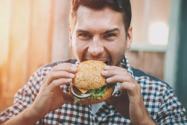 Hungriger Junger Mann Beißt Leckeren Hamburger Und Schaut Die Kamera Stockfoto