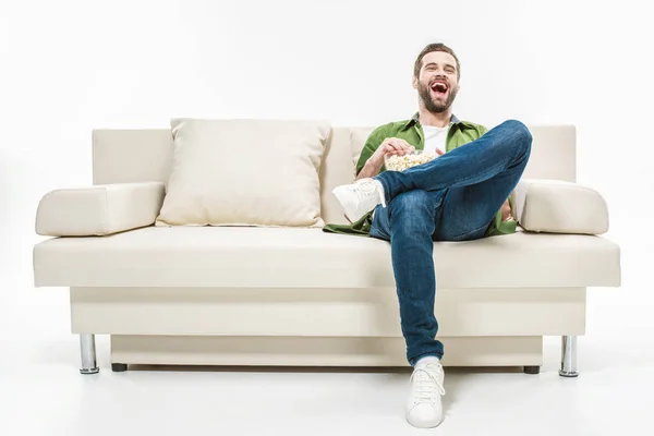 Rire jeune homme assis sur le canapé avec du pop-corn isolé sur blanc — Photo de stock
