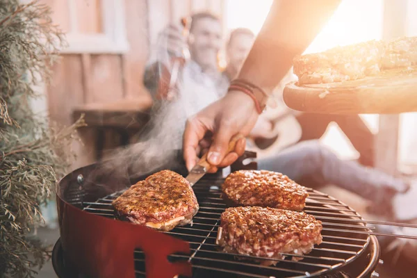 Tiro recortado de hombre haciendo barbacoa en el porche con luz de fondo y amigos borrosos en el fondo - foto de stock