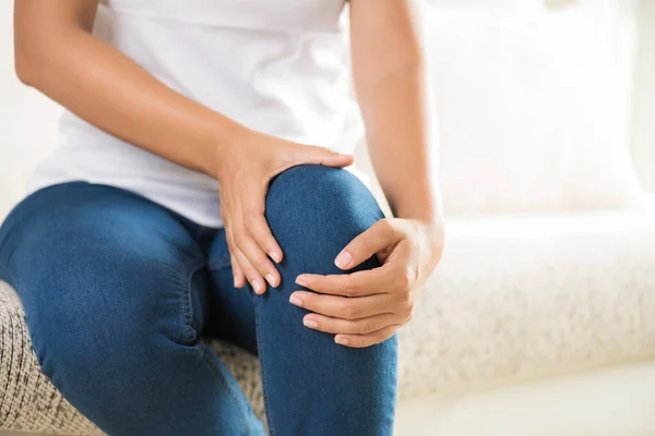 Closeup young woman sitting on sofa and feeling knee pain and she massage her knee at home. Healthcare and medical concept.