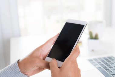 Ofiste bir dizüstü bilgisayar ile bir masada beyaz cep telefonu tutan kadın el.