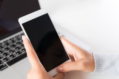 Ofiste bir dizüstü bilgisayar ile bir masada beyaz cep telefonu tutan kadın el.