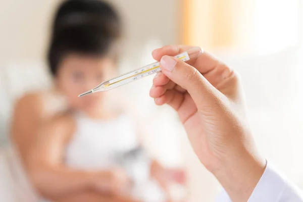 Doctor holding digital thermometer to measuring temperature of her ill kid and sick little girl with mother in background.