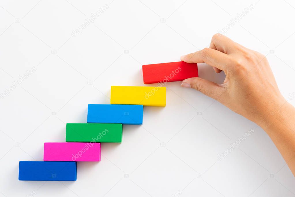 Concept of building success foundation. Women hand put red wooden block on colorful wooden blocks in the shape of a staircase.