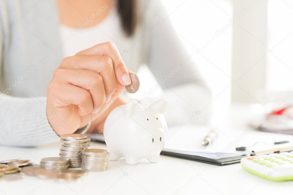 Woman hand putting money coin into piggy bank with stack of coins. Computer calculator on office table. Saving money wealth and financial concept.