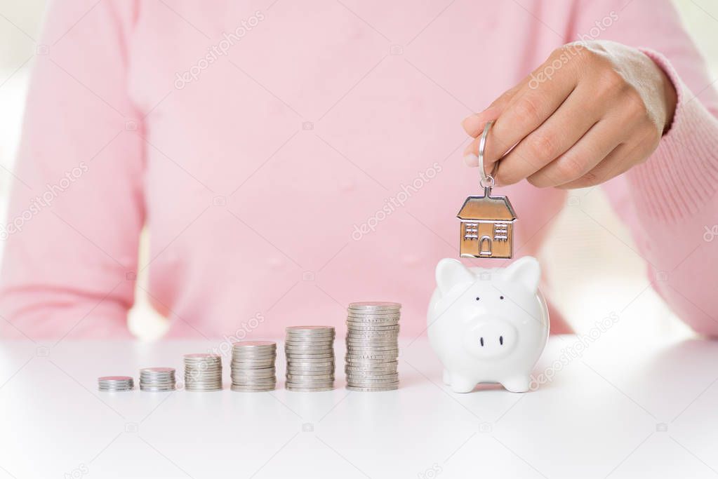 Woman hand holding house keychain with stack of coins and putting money bank note dollar into piggy bank . Saving money wealth for house and financial concept.