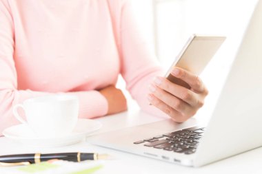 Ofiste bir dizüstü bilgisayar ile bir masada beyaz cep telefonu tutan kadın el.