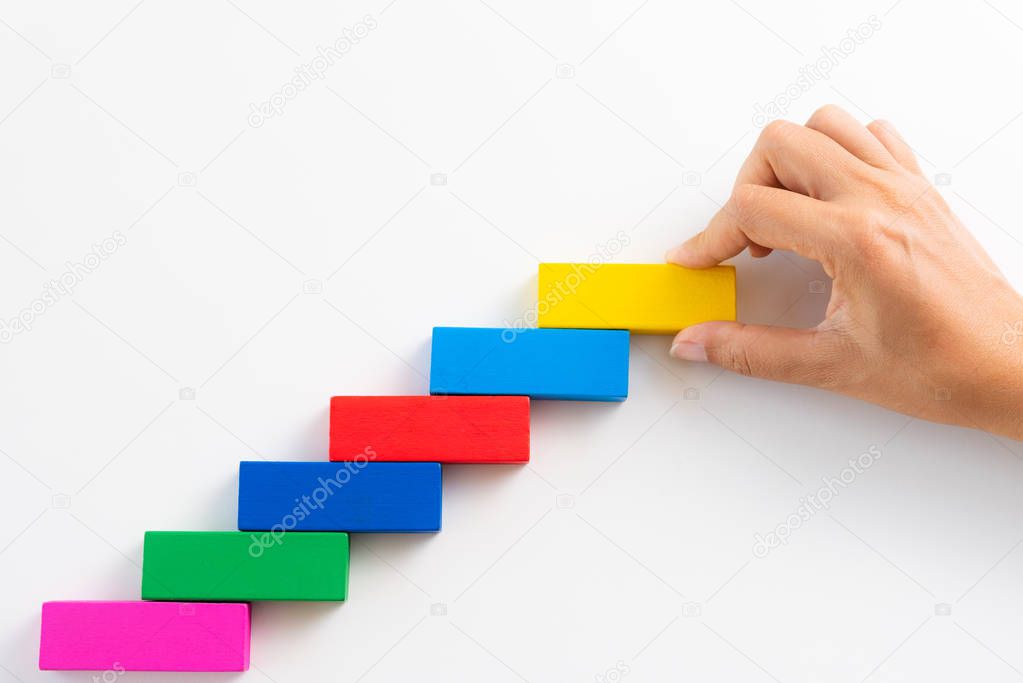 Concept of building success foundation. Women hand put red wooden block on colorful wooden blocks in the shape of a staircase.