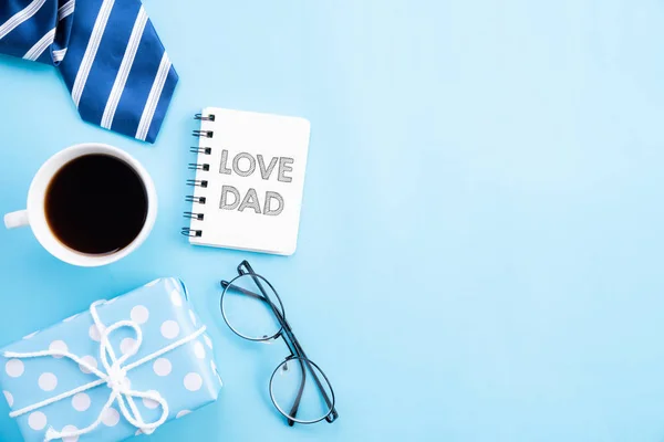 Concepto del día del padre feliz. Vista superior de corbata azul, hermosa caja de regalo, taza de café, vasos con texto LOVE DAD sobre fondo pastel azul brillante . — Foto de Stock