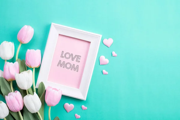 Happy mothers day concept. Top view of pink tulip flowers and white picture frame with LOVE MOM text on green pastel background. Flat lay. — Stock Photo, Image