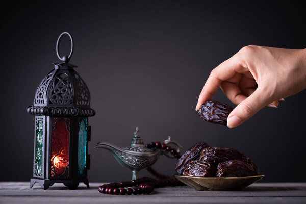 Ramadan food and drinks concept. Woman hand reaches out to a plate with date with Ramadan Lantern with arabian lamp, wood rosary, tea, dates fruit and lighting on a wooden table on dark background.