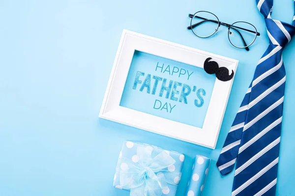 Happy fathers day concept. Top view of blue tie, beautiful gift box, white picture frame with Happy father's day text on bright blue pastel background. Flat lay.