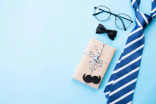 Happy fathers day concept. Top view of blue tie, beautiful gift box, glasses and mustache on bright blue pastel background. Flat lay.