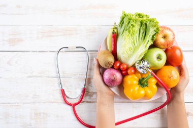 Sağlıklı yaşam tarzı, gıda ve beslenme kavramı. Beyaz ahşap masa üzerinde yatan stetoskop ile taze sebze ve meyve plaka tutan doktor kadın el kadar yakın.