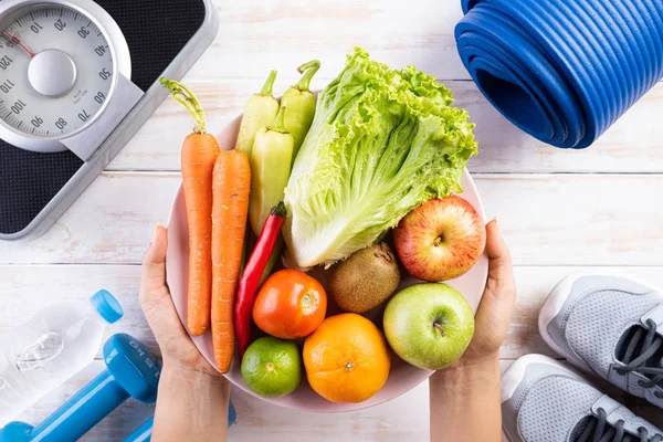 Estilo de vida saudável, comida e conceito de esporte. Visão superior da mulher mão segurando prato de legumes frescos e frutas com equipamento de atleta Balança de peso haltere azul, garrafas de água esporte no fundo de madeira branca . — Fotografia de Stock