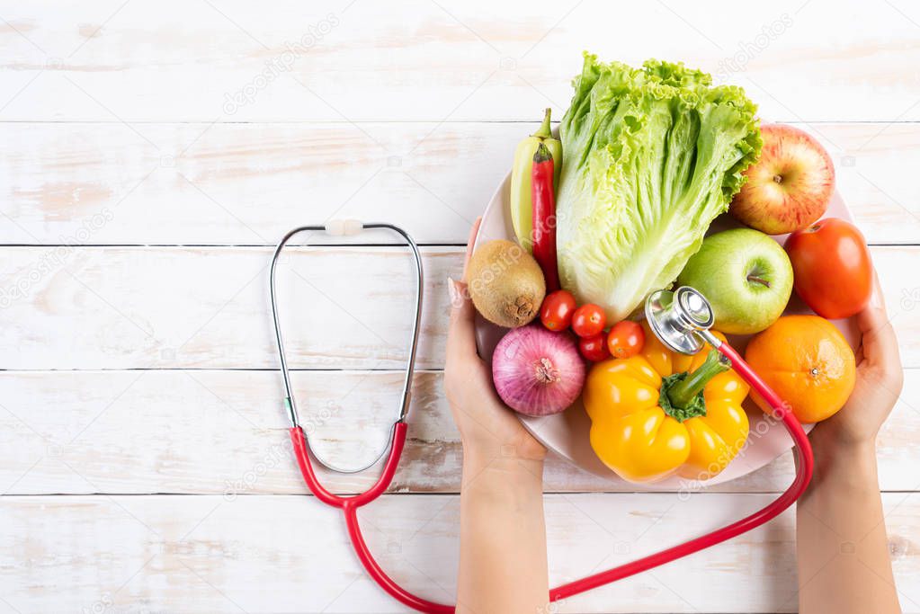 Healthy lifestyle, food and nutrition concept. Close up doctor woman hand holding plate of fresh vegetables and fruits with stethoscope lying on white wooden table.