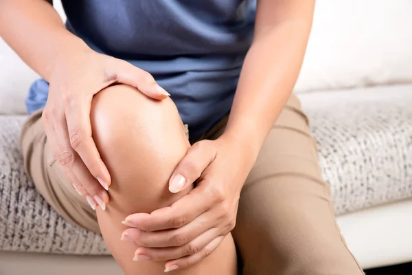 Close-up jovem mulher sentada no sofá e sentindo dor no joelho e ela massagear o joelho em casa. Conceito médico e de saúde . — Fotografia de Stock