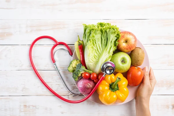 Sağlıklı yaşam tarzı, gıda ve beslenme kavramı. Beyaz ahşap masa üzerinde yatan stetoskop ile taze sebze ve meyve plaka tutan doktor kadın el kadar yakın. — Stok fotoğraf
