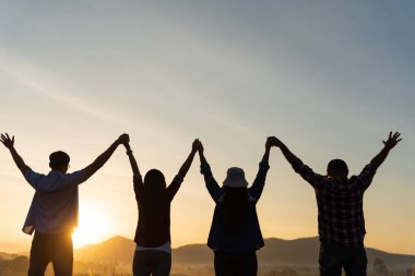 Bir grup mutlu arkadaş dağın önünde el ele tutuşarak eğleniyorlar ve birlik ve takım çalışmasını göstererek gün doğumunun tadını çıkarıyorlar. Arkadaşlık mutluluğu eğlence ortaklık takım konsepti.