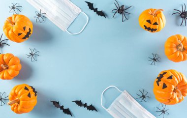 Cadılar Bayramı süslemeleri, pastel mavi arka planda balkabağı, kağıt yarasalar ve cerrahi maske. Covid-19 salgını sırasındaki Cadılar Bayramı kutlamalarının üst görüntüsü..