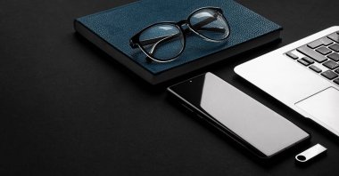 Siyah bir matte masa arkasında bir dizüstü bilgisayar, bir deri mavi defter, gözlük, bir kalem ve yeni bir akıllı telefon fotokopi alanı bulunur. 