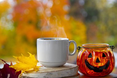 Bulanık bir sonbahar ormanının arka planında bir fincan sıcak kahve ve buharlı Cadılar Bayramı süslemeleri duruyor. 