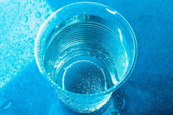 Одноразовое пластиковое стекло или чашка со свежей чистой водой на ярко-голубом фоне, вид сверху — стоковое фото