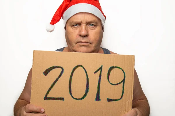 Funny senior serious man in red Santa Claus Christmas hat holds cardboard with 2019 numbers in hands and angrily looks at camera, humor Happy New Year concept